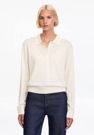 Mujer rubia con una camisa polo de manga larga crema y vaqueros azul oscuro, de pie frente a un fondo blanco.