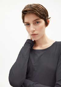 Langärmliges Oberteil aus dunkelgrauem Stoff mit rundem Ausschnitt und glatter Textur. Das Model hat kurze Haare und legt sanft die Hand auf den Hals.