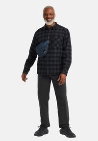 Geruite overhemd in zwart en grijs, gecombineerd met zwarte broek en donkere sportschoenen. Een blauwe schoudertas met ritszakken maakt de look compleet.