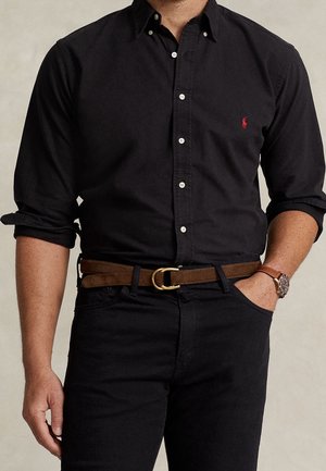 Mężczyzna ubrany w czarną koszulę na guziki z podwiniętymi rękawami, czarne spodnie, brązowy zamszowy pasek ze złotą klamrą oraz zegarek na brązowym skórzanym pasku.