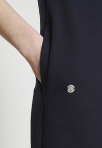 Marinfärgad klänning i slim fit, med en sidficka och en silverknapp med ingraverad detalj på framsidan. Slät textur.