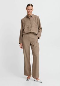 Camicia button-up marrone a maniche lunghe con due tasche frontali, abbinata a pantaloni su misura coordinati. Tessuto testurizzato e vestibilità rilassata. Tacchi argentati.