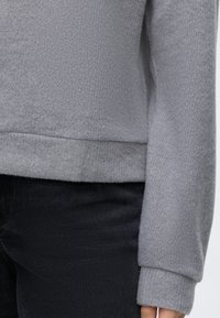 Personne portant un pull texturé gris clair à manches longues et un pantalon noir, visible de la taille jusqu'à mi-cuisse sur fond blanc.