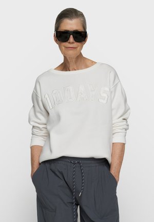 Ældre kvinde iført sorte store solbriller, hvid sweatshirt med teksten "10DAYS" og mørkegrå bukser med snørre, hænderne i lommerne.