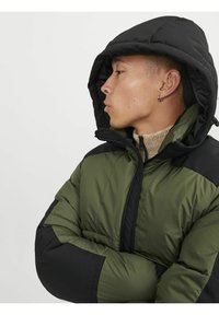 Giacca a vento verde e nera presenta un cappuccio, chiusura con zip, materiale testurizzato e un design color block a contrasto su spalle e maniche.