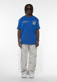 T-shirt blu in cotone con maniche corte, caratterizzata da testo grafico nella parte anteriore; abbinata a pantaloni larghi grigi chiari e sneakers bianche.