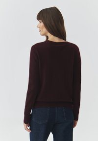 Maglione a maglia borgogna con scollatura rotonda, orlo a coste e maniche lunghe, indossato con jeans blu scuro. Texture morbida e design minimale.