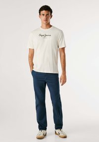 T-shirt blanche avec le logo "Pepe Jeans London", associé à un pantalon bleu marine et des baskets crème. Le mannequin se tient les mains dans les poches.