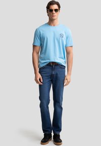 Hellblaues Baumwoll-T-Shirt mit rundem Halsausschnitt und seitlichem Logo, kombiniert mit dunkelblauen Jeans und schwarzen Turnschuhen. Die Accessoires umfassen eine Sonnenbrille.