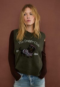 Olivgrön stickad väst med en dachshundgrafik i rosa och vitt, med texten "The Doggie Club" och "est. 1994". Ribbad nederkant.