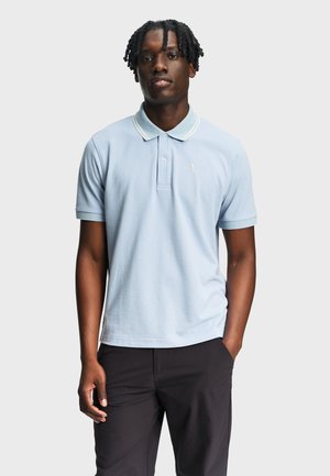 Mann mit kurzem, gedrehtem Haar, der ein hellblaues Polohemd mit kurzen Ärmeln und eine schwarze Hose trägt, mit einer Hand in der Tasche stehend.