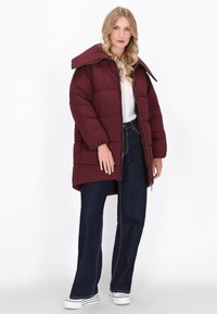 Giacca puffer bordeaux con un ampio colletto, design trapuntato e zip frontale, abbinata a jeans scuri e scarpe da ginnastica bianche.