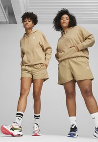 Puma beiges Hoodie und Shorts mit weißen Logos, kombiniert mit bunten Sportschuhen und gestreiften Socken, die einen lässigen Schnitt und weichen Stoff aufweisen.