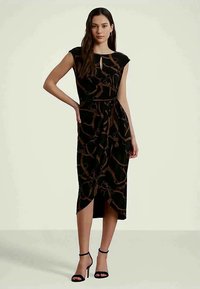 Sieviete, kura ģērbjas bez piedurknēm melnā midi kleitā ar brūnu ķēdes printu, ar atslēgas caurumu pie kakla, sasietu jostu un melniem potīšu siksniņu apaviem, stāv uz viendabīga fona.