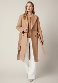 Długi płaszcz wełniany w kolorze camel z wyraźnymi klapami, podwójnym zapięciem z przodu i dwoma bocznymi kieszeniami, noszony na lekkim swetrze i białych szerokich spodniach.