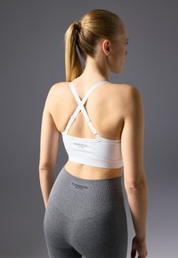 Weißer Sport-BH mit X-Back-Trägern, der eine glatte Textur und dezente Markenkennzeichnung aufweist. Kombiniert mit hochgeschnittenen grauen Leggings mit gerippten Akzenten.