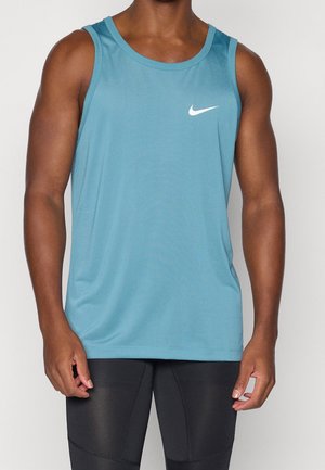 Athletische Person, die ein blaues ärmelloses Nike-Oberteil und schwarze enganliegende Sporthosen vor einem schlichten weißen Hintergrund trägt.