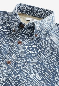 Marineblaue Hemd mit weißen geometrischen Mustern. Verfügt über eine Knopfleiste mit braunen Knöpfen und einem strukturierten inneren Kragenfutter.
