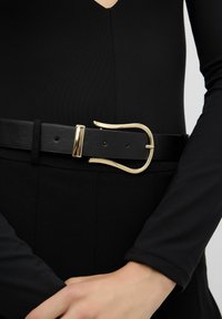 Cinturón de cuero negro con una correa ancha y un distintivo hebilla dorada de diseño curvado, usado sobre una prenda negra.