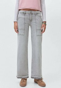 Pantaloni cargo in denim grigio a gamba larga con tasche frontali, dettagli con orlo grezzo e una vestibilità rilassata. Abbinati a calzature beige per un contrasto.