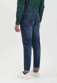 Jeans in denim blu scuro con una vestibilità comoda, dotati di polsini arrotolati, due tasche posteriori e una patch in pelle marrone nella zona della vita.