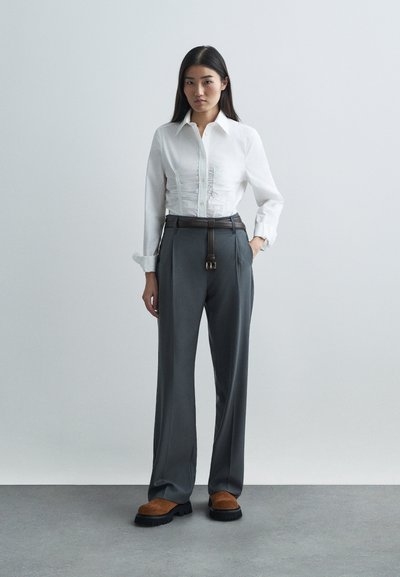 Bjela košulja s naborima sprijeda, u paru s grafitnim krojenim hlačama i smeškim semiš cipelama na potplatima deblje crne boje.