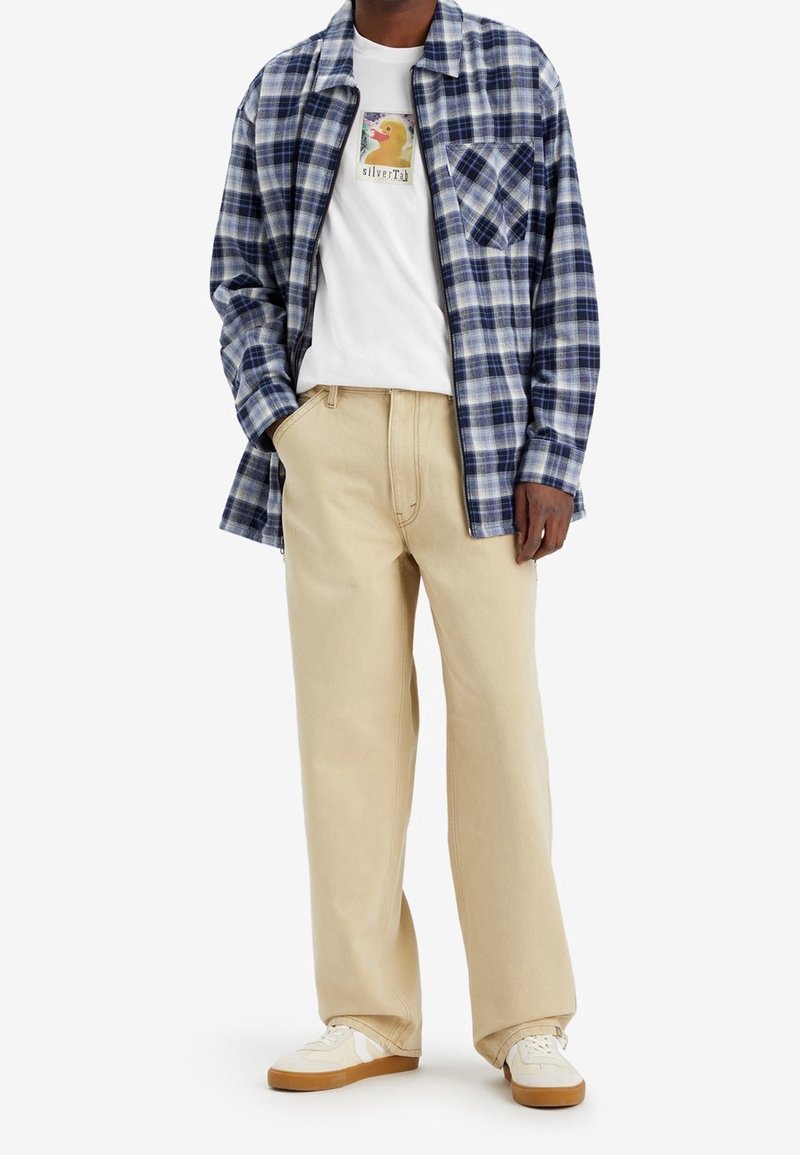 El atuendo masculino incluye una camisa de cuadros azul y blanca, una camiseta gráfica blanca, pantalones beiges y zapatillas blancas con suela de goma.