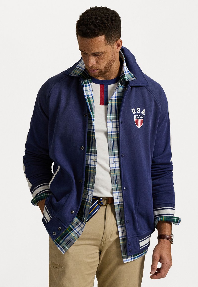 Mand iført en lagdelt blå varsityjakke med USA-emblem, grønternet skjorte, hvid t-shirt, beige bukser og stribebælte, hænderne i lommen.