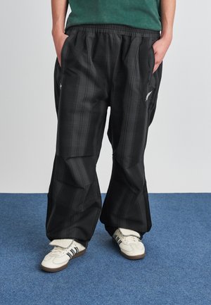 Personne portant un pantalon ample à carreaux noirs et des baskets blanches avec des rayures noires, debout sur un tapis bleu.