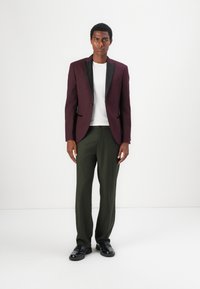 Bordeauxfarbener Blazer mit schwarzen Revers, darunter ein weißes T-Shirt, olivgrüne Hosen und schwarze Schuhe. Schlanker Schnitt und glatte Textur.