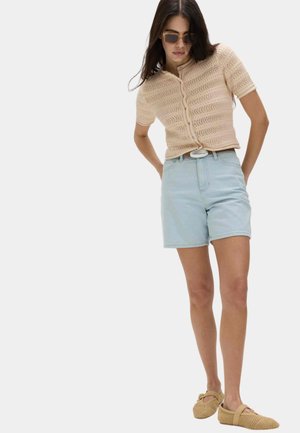 Frau trägt beiges gestricktes Kurzarmoberteil, hellblaue hoch taillierte Denim-Shorts, Sonnenbrille und beige gewebte flache Schuhe, steht mit den Händen hinter dem Rücken.