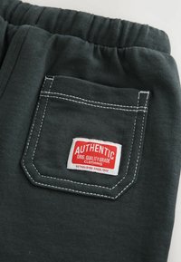 Pantalons de survêtement en tissu gris avec une taille élastique et une poche arrière ornée d'une étiquette "AUTHENTIC" rouge et blanche cousue.
