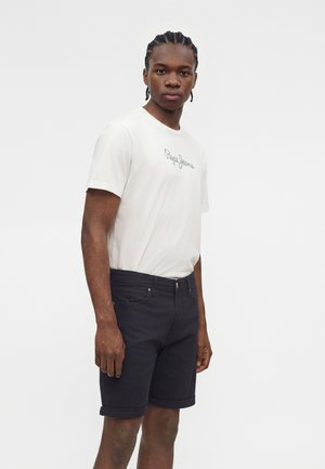 Άνδρας με πλεξούδες που φοράει λευκό μπλουζάκι "Pepe Jeans" και μαύρα σορτς με γυρισμένα ρεβέρ, στέκεται μπροστά σε λευκό φόντο.