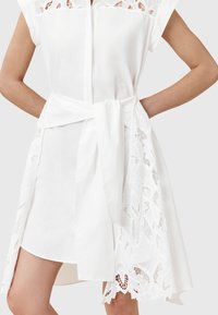 Weißes Kleid mit kurzen Ärmeln, Spitzenakzenten am Saum und einer gebundenen Taillendetails. Glattes Material mit einem leichten transparenten Effekt.