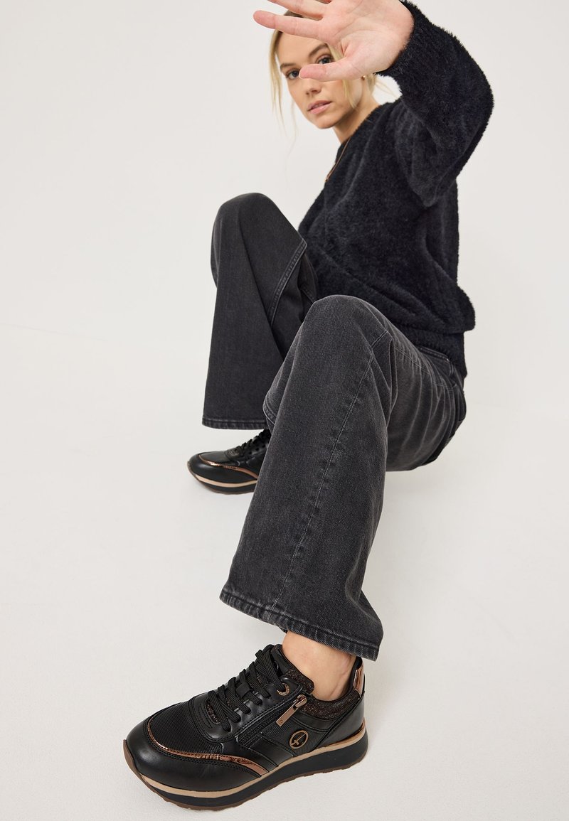 Black sneakers with a glossy leather upper, textured accents, and bronze stitching. Thick sole and lace-up design, showing a casual look.