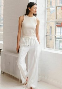 Lichtbeige mouwloze top in combinatie met witte wijde broek. De broek heeft een elastische tailleband en zijzakken, met een soepele textuur.