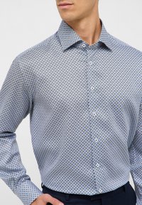 Camicia con bottoni in tessuto leggero; presenta un motivo geometrico blu, bottoni bianchi e un colletto strutturato. Forma aderente, maniche lunghe.