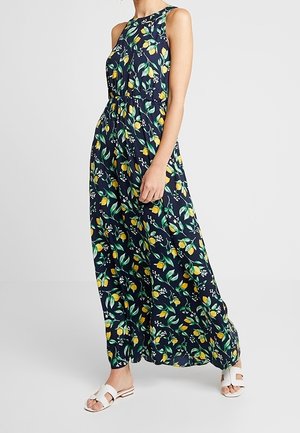 Frau trägt ein ärmelloses marineblaues Maxikleid mit gelben Blumen- und grünen Blattmustern, kombiniert mit weißen flachen Sandalen.