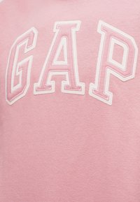 Ροζ ύφασμα με μεγάλα λευκά και ροζ κεντημένα γράμματα που σχηματίζουν τη λέξη "GAP" στο κέντρο της επιφάνειας.