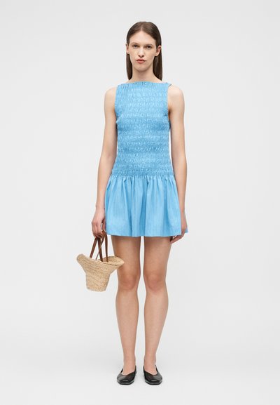 Γυναίκα που φοράει ένα μπλε κοντό φόρεμα χωρίς μανίκια με βολάν, κρατώντας μια μικρή υφαντή τσάντα από άχυρο και φοράει μαύρα ίσια παπούτσια, στέκεται όρθια.