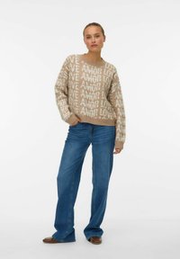 Femme debout les mains dans les poches, portant un pull beige avec le texte "LOVE AMOR" et un jean large bleu, sur fond blanc.