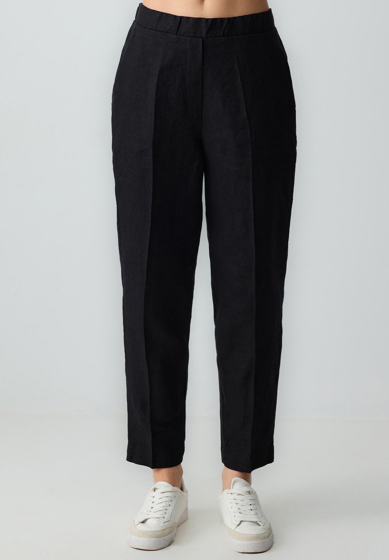Schwarze Leinenhose mit taillierter Passform, vorderen Falten, schmal zulaufenden Beinen und flachem Bund, kombiniert mit weißen Sneakers.