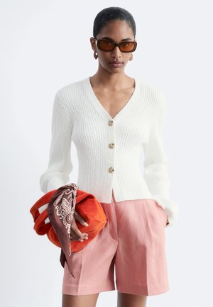 Femme portant un cardigan blanc côtelé boutonné, un short rose, des lunettes de soleil marron, tenant un sac orange avec une écharpe marron à motif.