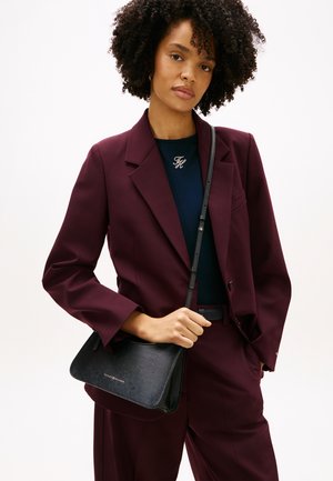 Γυναίκα με σγουρά μαλλιά που φοράει μια μπορντό κοστούμι και μπλε τοπ, κρατώντας μια μαύρη τσάντα χιαστί με χρυσά γράμματα.