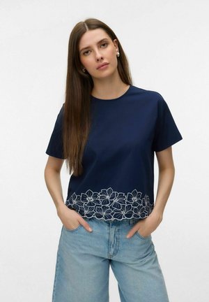 Donna con capelli lunghi castani che indossa una maglietta blu navy a maniche corte con ricamo floreale bianco sull'orlo e jeans azzurri, con le mani in tasca.