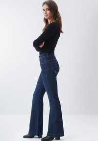 Jeans acampanados de mezclilla azul oscuro deslavado con cintura alta, bolsillos traseros cosidos y dobladillo limpio, combinados con una camiseta negra de manga larga.