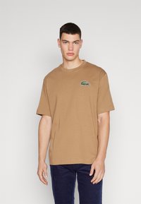 T-shirt in cotone marrone con maniche corte e scollatura a girocollo, con un piccolo logo ricamato verde sul lato sinistro del petto.