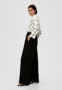 Witte blouse met zwart abstract patroon, gecombineerd met losse zwarte broek. De outfit heeft een ontspannen pasvorm en zakken op de broek.