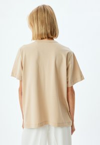 Beige lyhythihainen t-paita, jossa on pyöreä kaula-aukko, valmistettu pehmeästä puuvillasta. Näkyvä ompelutaito; rento malli. Takakatsaus, ei graafisia kuvastoja tai kuvioita.