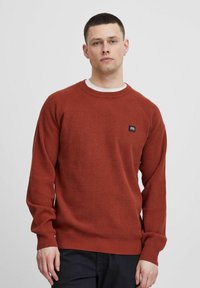 Maglione color ruggine con lavorazione a maglia testurizzata, scollatura a girocollo e maniche raglan. Presenta un piccolo etichetta del marchio nera sul petto.